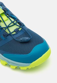Modra tekaška čevlja z teksturirano mrežasto zgornjico, živahnimi zelenimi akcenti, varnim sistemom vezalk in svetlo rumenim podplatom za oprijem.