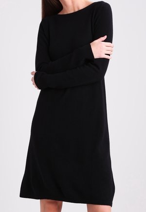 Kobieta ubrana w czarną sukienkę z długim rękawem sięgającą do kolan, z okrągłym dekoltem, stojąca na jednolitym jasnym tle.