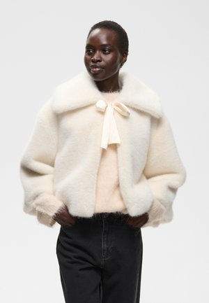 Femme portant un manteau blanc duveteux avec un grand col et un nœud papillon, associé à un pantalon noir, debout les mains dans les poches.