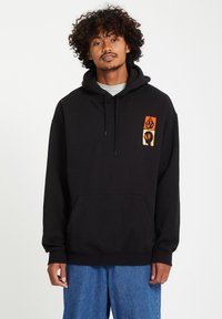 Zwarte hoodie met een trekkoord, voorzien van een voorzak en oranje grafische ontwerpen op de linker borst. Gedragen over een grijze shirt.