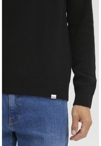 Man wearing a black long-sleeve sweater and blue denim jeans, hand relaxed by side, sweater label visible near hem.