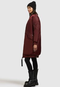 Bordeaux parka met een gladde afwerking, voorzien van een rits aan de voorkant, zijzakken en een geribbelde zoom. Geddragen met zwarte leggings en combat boots.