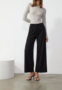 Haut gris à manches longues avec col rond, associé à un pantalon noir taille haute à jambes larges. Le mannequin porte des talons noirs à brides. Texture lisse.