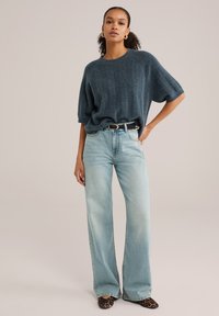 Suéter de punto en un tono verde azulado oscuro y de gran tamaño, con textura acanalada, combinado con jeans de pierna ancha en un tono de lavado claro y zapatos de estampado de leopardo. Se ve un cinturón negro.