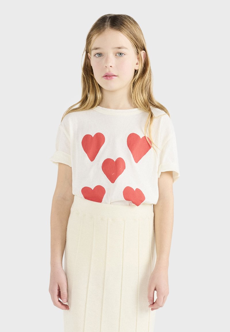 Λευκό βαμβακερό t-shirt με μεγάλες κόκκινες καρδιές. Συνδυασμένο με κρεμ φούστα με πτυχές. Απλός, καθημερινός σχεδιασμός.