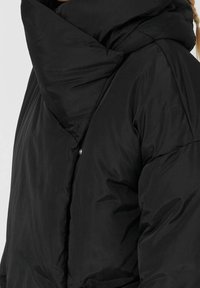 Sort pufferjakke med høj krave, geometrisk design, glat tekstur og en knapdetalje på forsiden.