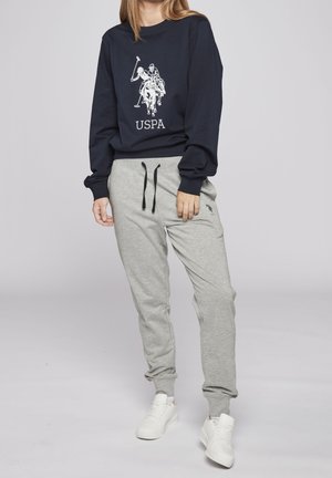 Person, der einen marineblauen Sweatshirt mit einem Polo-Spieler-Logo und graue Jogginghose mit schwarzem Kordelzug sowie weißen Sneakers trägt und steht.