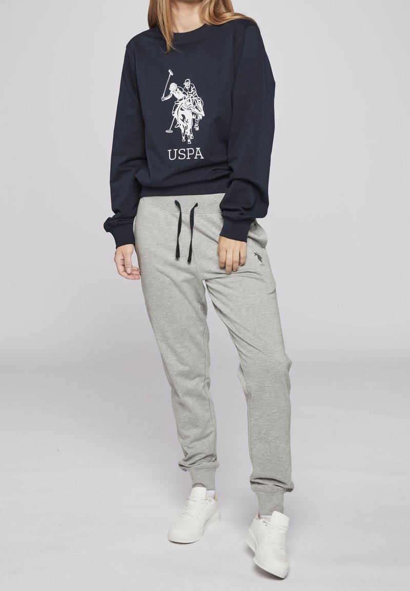 Osoba oblečená v tmavě modrém svršku s logem pólisty a šedých teplácích s černou šňůrkou a bílými teniskami, stojící.