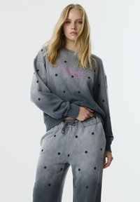 Grijze sweatshirt met zwarte stippen, voorzien van een logo in het roze. Gecombineerd met bijpassende joggingbroek, ook met stippen en een trekkoord in de taille.