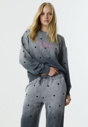 Felpa grigia con pois neri, dotata di un logo rosa. Abbbinata a pantaloni della tuta coordinati, anch'essi con pois e vita con cordino.