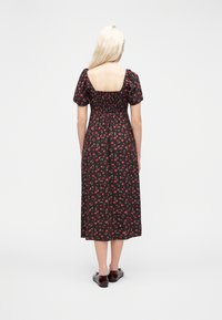 Γυναίκα με μακριά ξανθά μαλλιά φοράει μαύρο μίντι φόρεμα με κόκκινο floral μοτίβο, φουσκωτά μανίκια, μαζεμένη πλάτη και σκούρα επίπεδα παπούτσια, στέκεται σε λευκό φόντο.