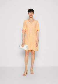 Robe à manches courtes et décolleté en V, de couleur orange clair avec un petit motif floral. Le modèle tient une pochette blanche et porte des sandales à talons beiges à lanières.