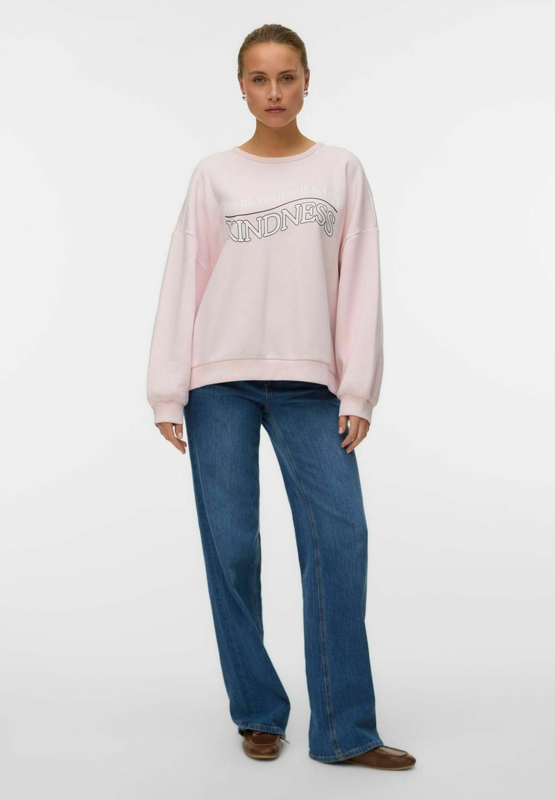 Sweatshirt oversize rosa chiaro con la scritta "Kindness" stampata in grigio. Abbinato a jeans blu a gamba larga e scarpe flat marroni.