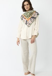 Grote sjaal met een patroon in tinten groen, roze en oranje die over een beige blouse is gedrapeerd. Model draagt lichtgrijze gebreide broek met wijde pijpen.
