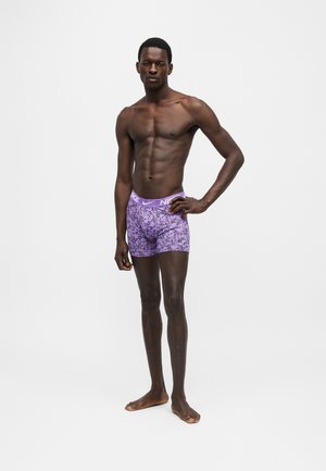 Αθλητικός άντρας στέκεται ξυπόλητος φορώντας μοβ μποξέρ Nike με μαύρο αφηρημένο σχέδιο και κορδόνι με λογότυπο.