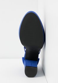 Chaussure à talon haut en daim bleu vue de la semelle avec un dessous noir texturé et un talon carré sur fond blanc en coin.