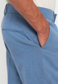 Pantalons ajustés bleu clair avec une texture lisse, dotés de poches latérales et d'un design épuré et simple. Coutures visibles et taille plate.