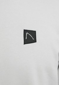 Camiseta de cuello redondo en gris claro, fabricada con tejido texturizado, que presenta un parche cuadrado negro con un diseño de líneas blancas, bordes cosidos y un acabado suave.