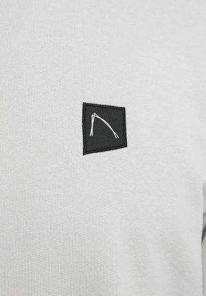 Camiseta de cuello redondo en gris claro, fabricada con tejido texturizado, que presenta un parche cuadrado negro con un diseño de líneas blancas, bordes cosidos y un acabado suave.