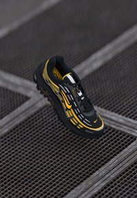 Zwarte en gele sport sneaker met veters en reflecterende details, boven een metalen rastervloer zwevend.