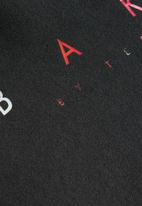 Tessuto nero con grandi elementi testuali in rosso e bianco. La superficie ha un aspetto texturizzato che mette in risalto i dettagli del design stampato.