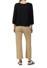 Persoon in zwarte lange mouwen top, beige rechte broek met rafelige zoom en witte sneakers met zwarte strepen, staand met de rug naar de camera.