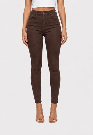 Woman wearing high-waist dark brown skinny jeans and clear open-toe heels, standing against a plain light background.