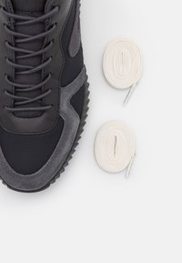 Baskets noires et grises avec une tige texturée, présentant une finition en cuir lisse et en suède. Deux lacets blancs sont placés à proximité.