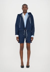 Denimjakke med hætte, frontlommer og snøre, parret med matchende denimshorts. Lyseblå skjorte under. Sorte sko.
