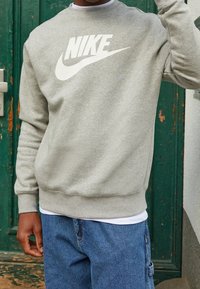 Sudadera gris con un gran logo blanco de Nike, puños y dobladillo acanalados, textura suave, usada sobre una camiseta blanca y combinada con jeans azules.
