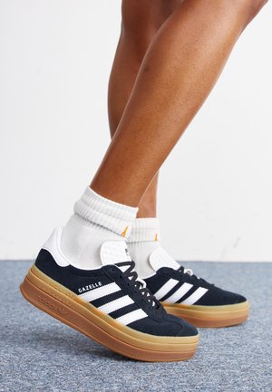 Ben iført hvide sokker og sorte Adidas Gazelle sneakers med hvide striber og tykke brune såler på en blå tæppebelagt gulv.