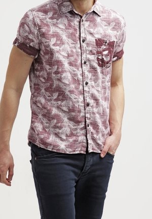 Homme portant une chemise à manches courtes boutonnée avec un motif floral bordeaux et blanc, associée à un jean bleu foncé, main dans la poche.