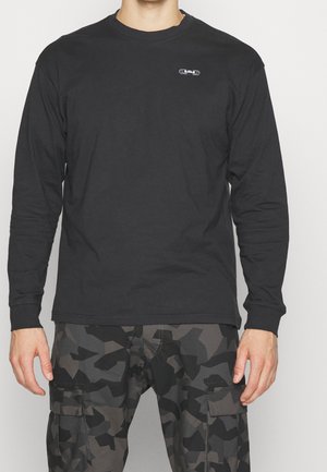 Mężczyzna ubrany w czarną koszulę z długim rękawem z małym logo na piersi oraz czarno-szare kamuflażowe spodnie bojówki na jednolitym tle.