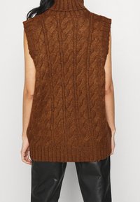 Hnědý pletený svetr bez rukávů s rolákem a vzorem copánků, s žebrovaným lemem, vyrobený z tkané příze, spárovaný s černými koženými kalhotami.