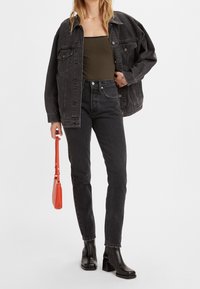 Mujer vestida con una blusa a rayas y una chaqueta de denim negra de gran tamaño, combinada con jeans negros y botines de tacón, sosteniendo un bolso pequeño rojo.