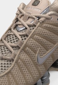 Μπεζ αθλητικό παπούτσι Nike Shox με πλέγμα και υφή στο επάνω μέρος, κορδόνια στο μπροστινό μέρος, ορατό ασημί λογότυπο Nike swoosh και αποσβεστήρες στη σόλα.