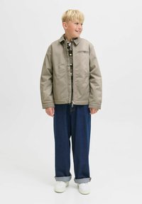Blond pojke som bär en beige jacka över en rutig skjorta, lösa mörkblå jeans med uppvikte ben, och vita sneakers, ser åt sidan.