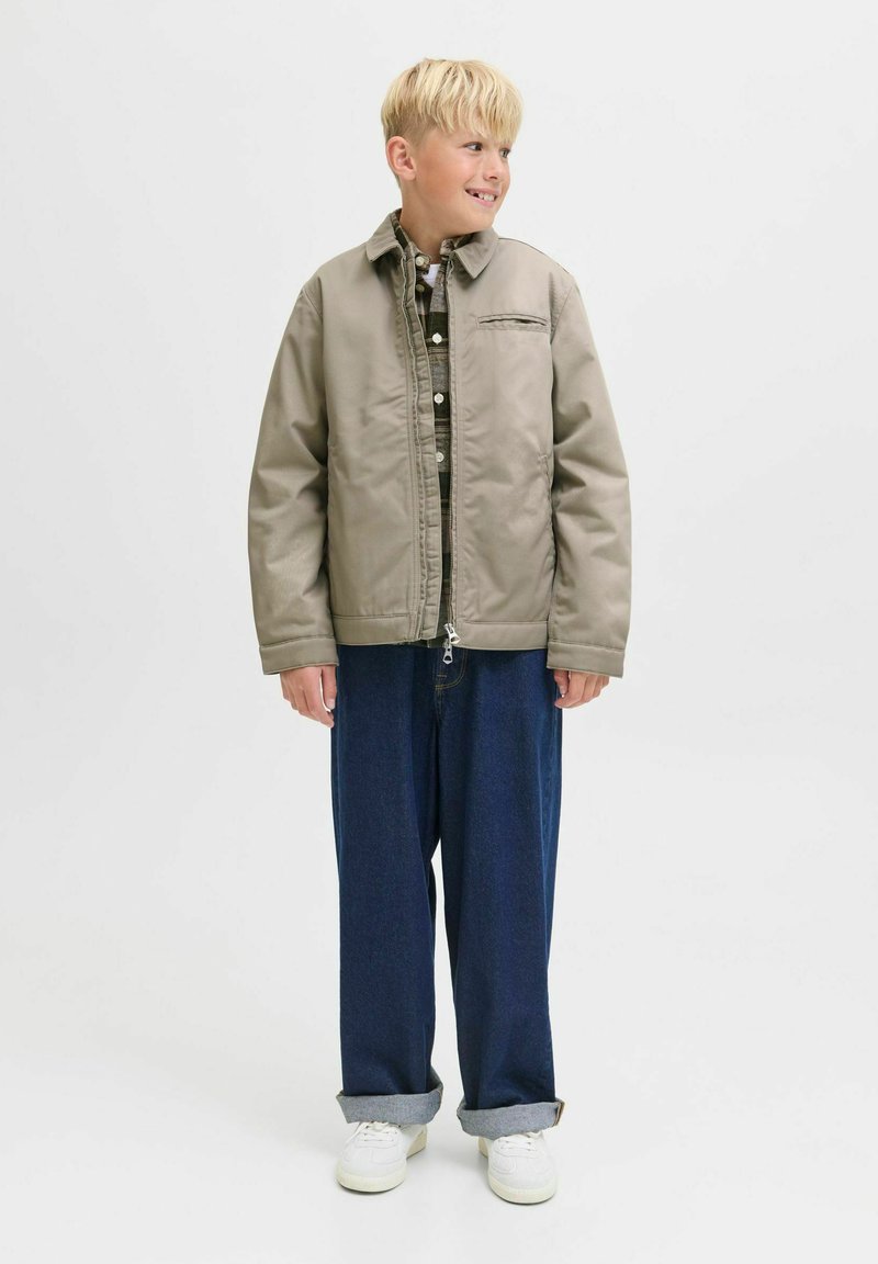 Blond pojke som bär en beige jacka över en rutig skjorta, lösa mörkblå jeans med uppvikte ben, och vita sneakers, ser åt sidan.