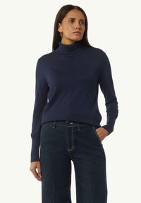 Granatowy sweter z golfem wykonany z gładkiego materiału z długimi rękawami, z subtelnym detalem szwu. Połączony z ciemnoniebieskimi spodniami dżinsowymi.