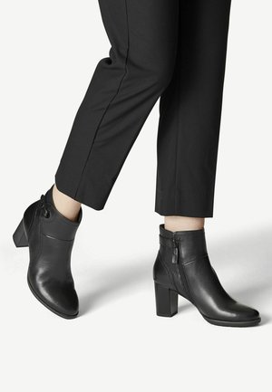 Bottines en cuir noir à talons blocs portées avec un pantalon noir court sur un fond clair.