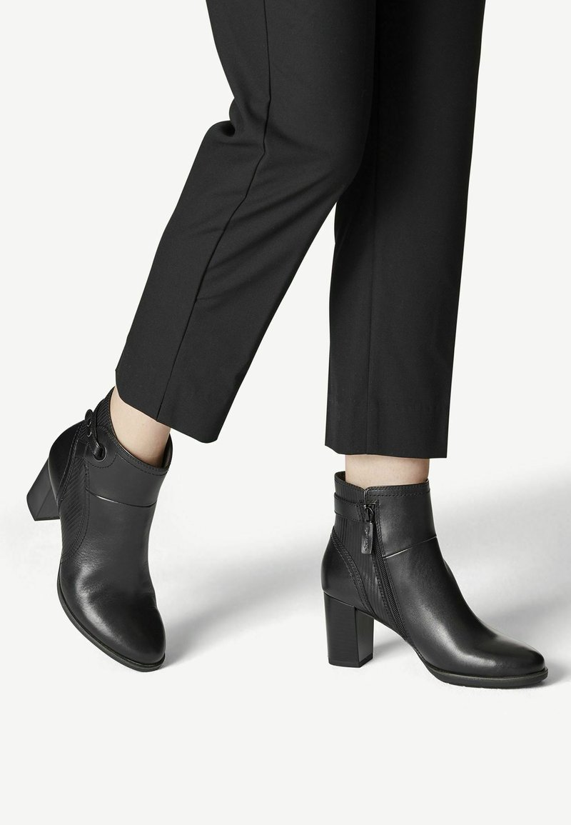 Bottines en cuir noir à talons blocs portées avec un pantalon noir court sur un fond clair.