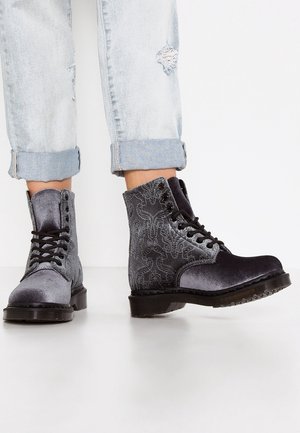Bottes de combat en velours gris usé avec des lacets noirs et des motifs décoratifs, assorties à un jean déchiré bleu clair retroussé.