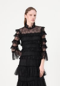 Svart spetsklänning med genomskinliga långa ärmar, hög krage och lager av volanger. Elegant textur med intrikata spetsmönster genom hela klänningen.