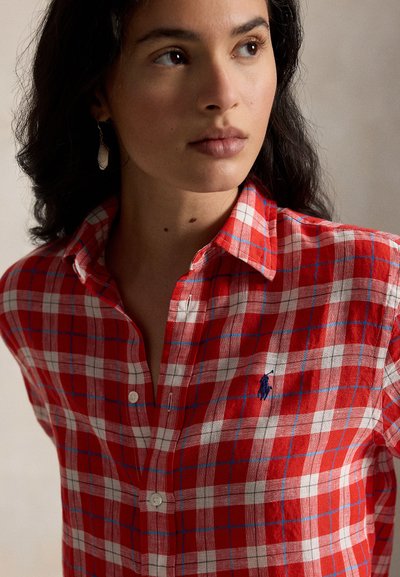 Kvinne med mørkt hår iført rød- og hvitrutete skjorte med blå logo, som ser litt til siden mot en ensfarget bakgrunn.