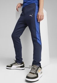 Marineblaue Jogginghosen mit einer taillierten Passform, die ein PUMA-Logo an der Hüfte und strukturierte Seitenpaneele aufweisen, kombiniert mit grauen und schwarzen Sneakers.