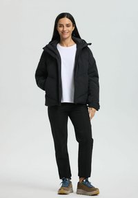 Eine lächelnde Frau, die eine schwarze Pufferjacke, ein weißes Hemd, schwarze Hosen und braune Wanderschuhe mit blauen Schnürsenkeln trägt, steht vor einem weißen Hintergrund.