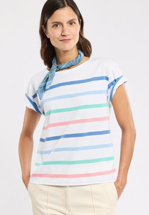 Femme aux cheveux bruns portant un t-shirt blanc avec des rayures horizontales multicolores et un foulard à motifs bleu autour du cou, les mains dans les poches d'un pantalon beige.