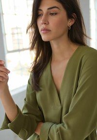Chemisier vert olive avec un décolleté en V et des manches longues et fluides. Fabriqué en tissu doux, il possède une seule poche sur la poitrine pour un ajout de détail.
