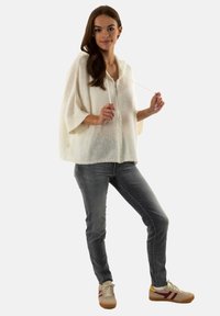 Cárdigan blanco de punto con un corte relajado y mangas anchas, que cuenta con una cremallera frontal. Combinado con jeans grises y zapatillas beige.