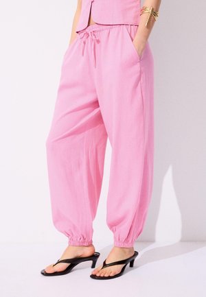 Osoba nosząca luźne różowe spodnie ze sznurkiem i elastycznymi mankietami, czarne sandały na obcasie z paskiem między palcami oraz złotą bransoletkę mankietową na prawej ręce.